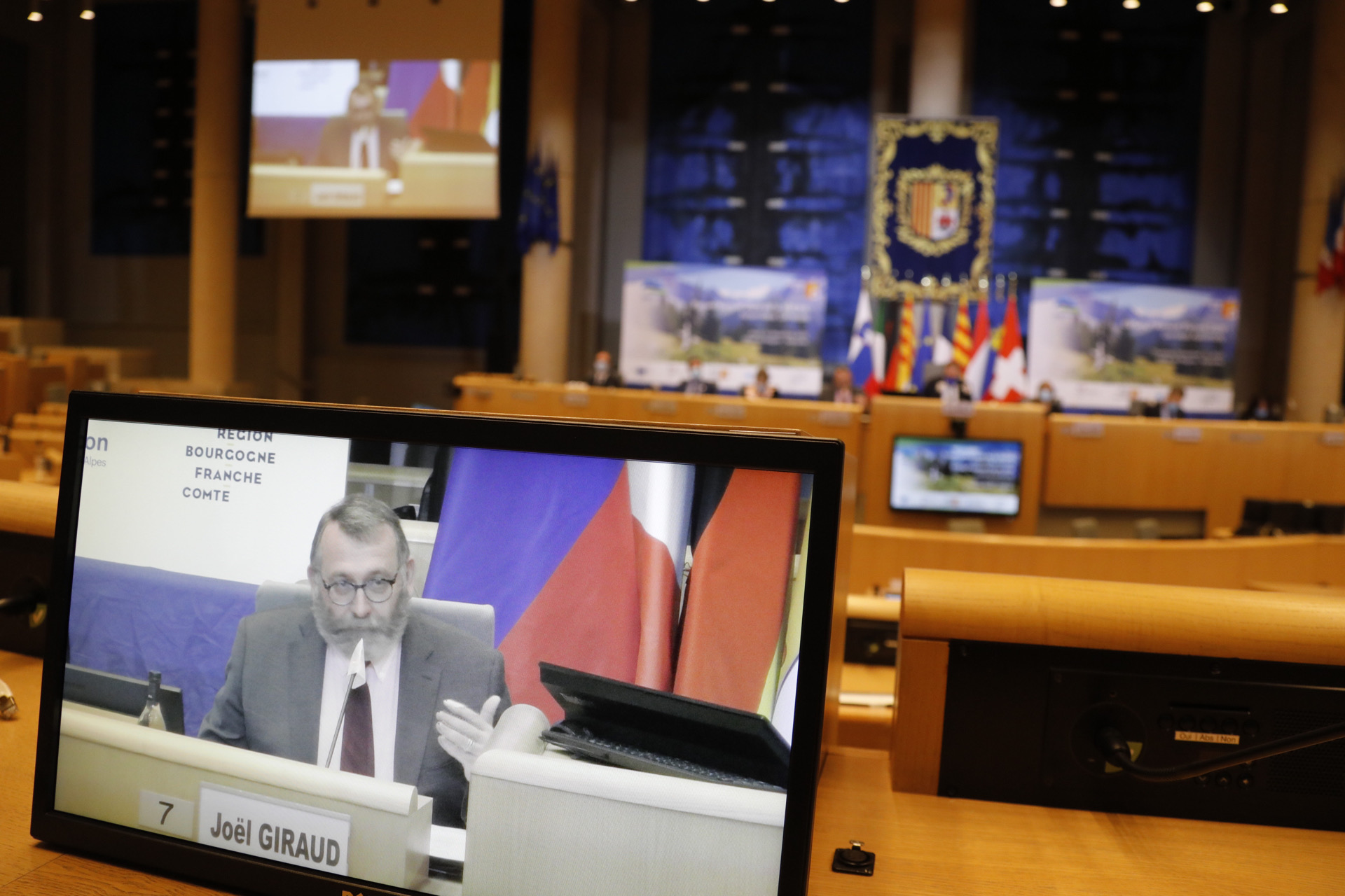 Photo du ministre Joël Giraud en train de parler à la tribune, vu à travers un écran de projection, avec l'hémicyle en arrière-plan.
