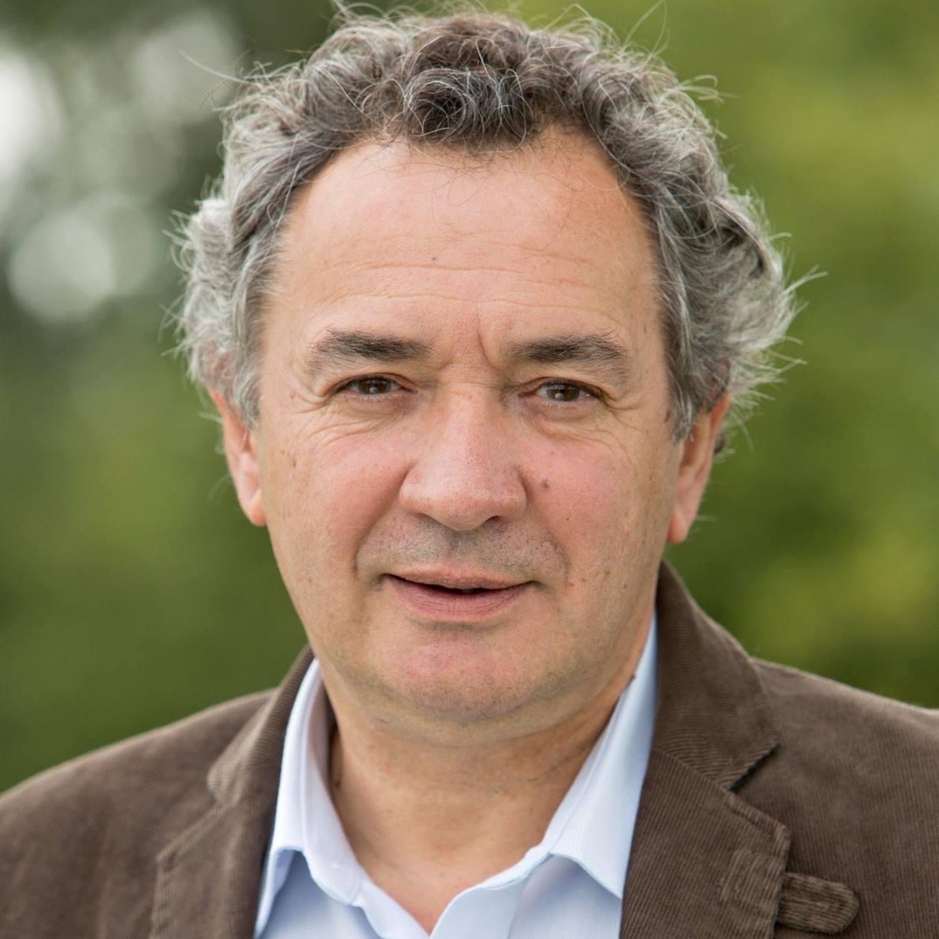 Photo portrait de Pierre Jarlier, cheveux gris, chemise blanche et veste marron