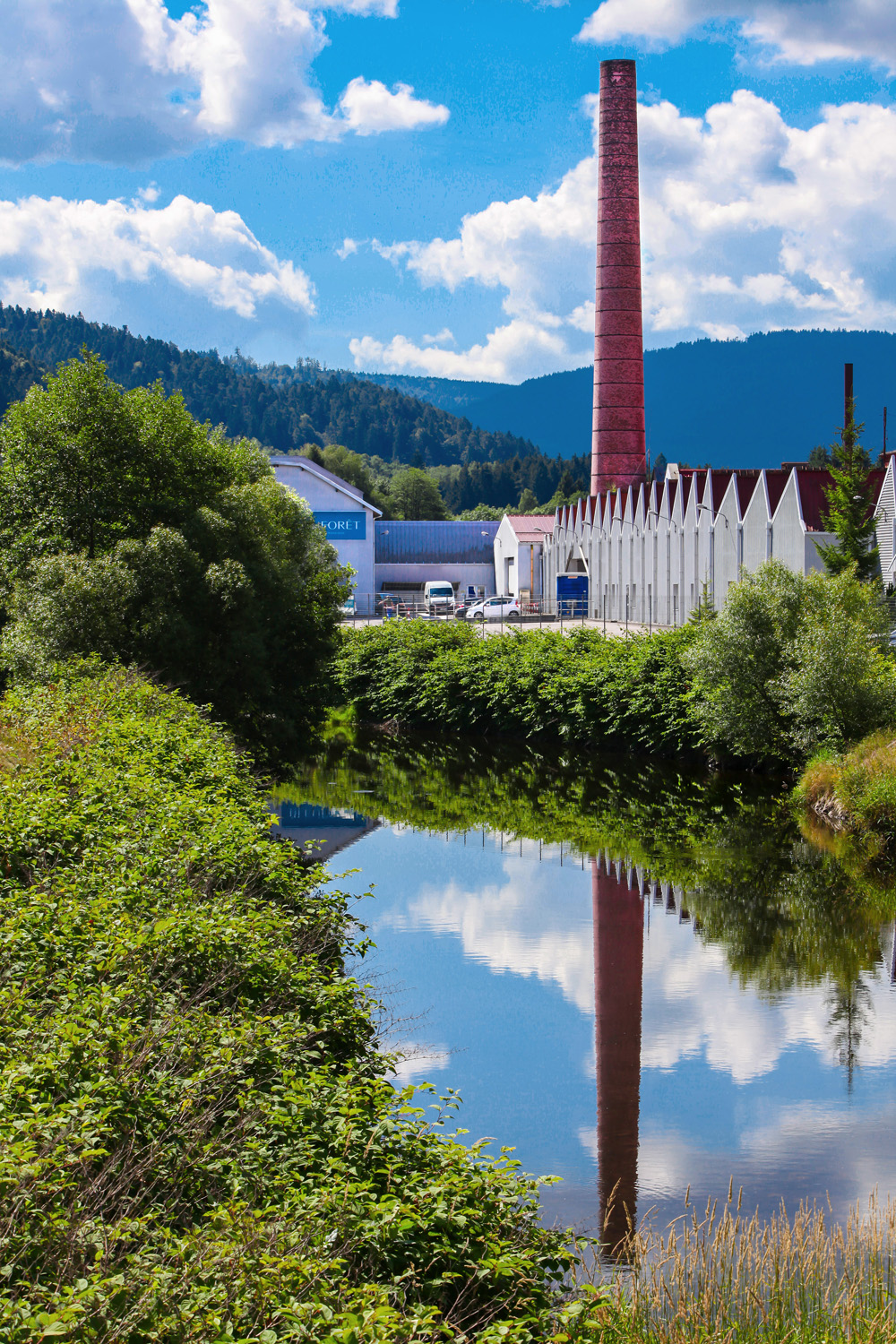 Vue sur une usine avec une grande cheminée en briques rouges, située le long d'une petite rivière et dans un cadre verdoyant, dans les Vosges