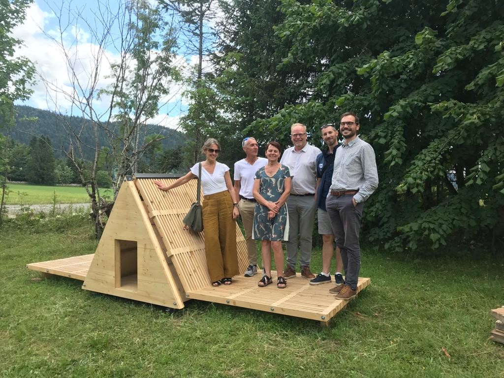 Un groupe de personnes pose à côté du prototype de cabane en bois, à taille réduite. Ils sont dans un espace vert, avc une colline en fond.