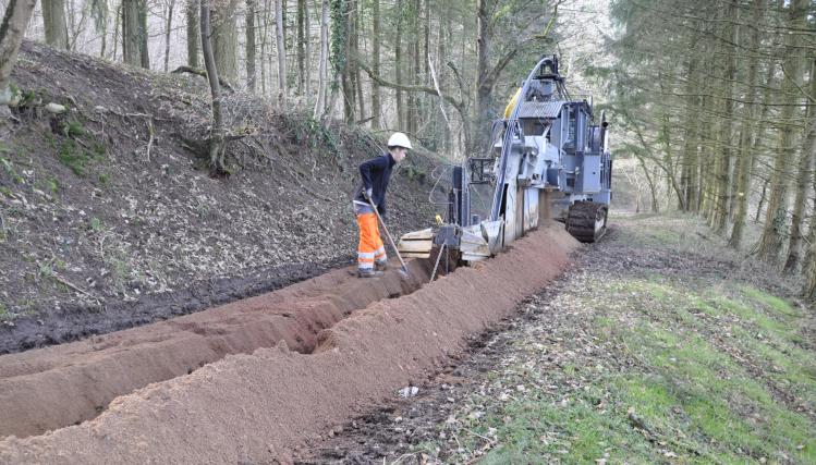 Installation de la fibre avec une machine qui creuse un fossé et un ouvrier qui déplpoie la ligne, dans un sous-bois