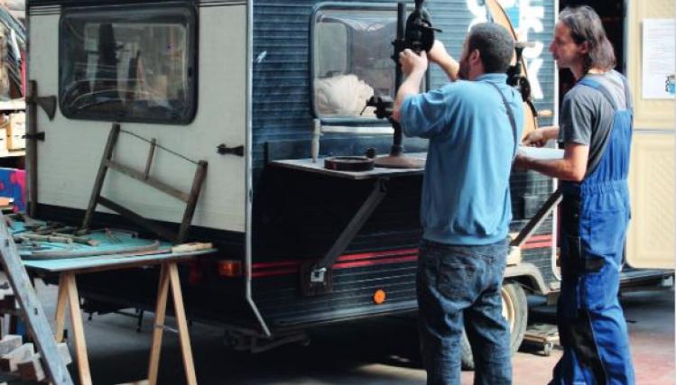 Deux hommes sont en train de réparer une vieille caravane blanche et bleu, dans un atelier. 