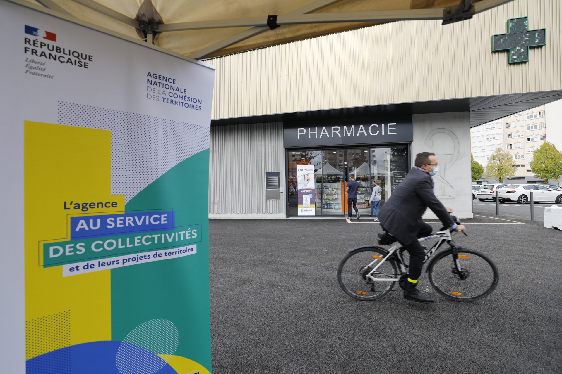 Kakémono de l'ANCT devant le centre comercial, avec un monsieur à vélo qui passe devant la nouvelle pharmacie