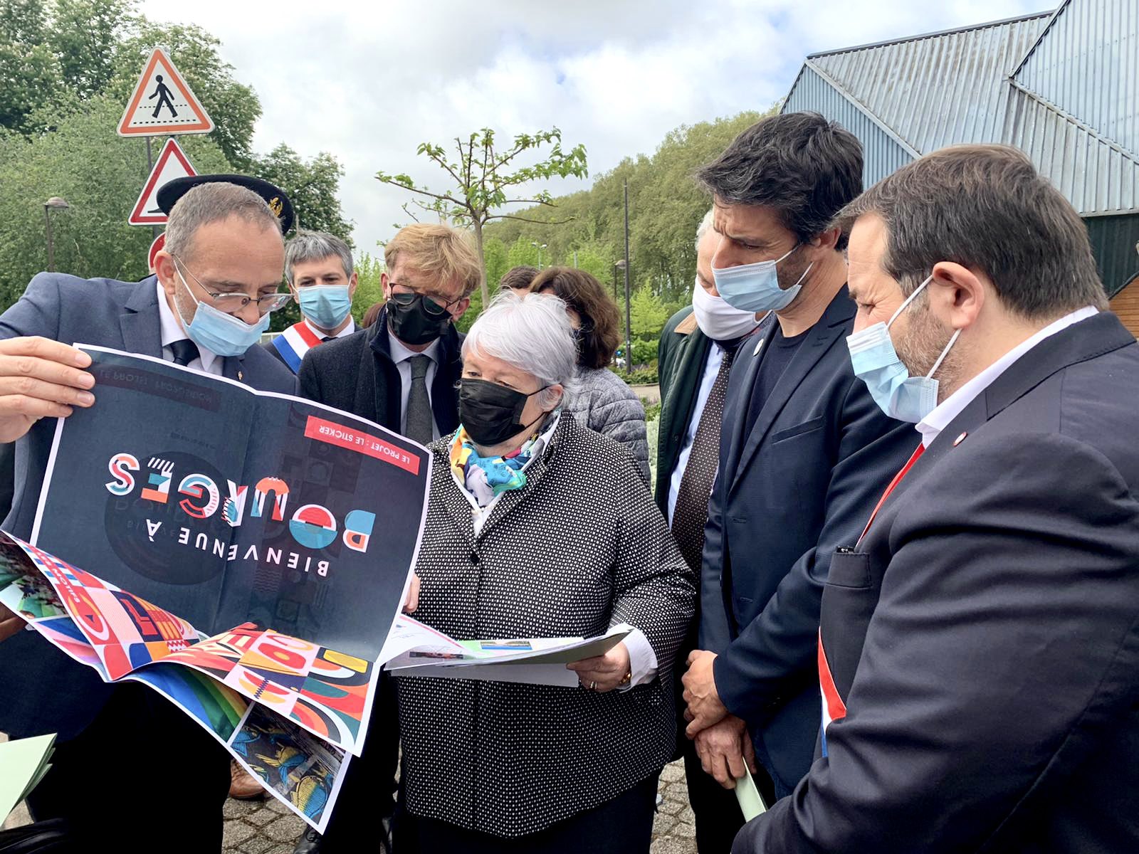 En extérieur, groupe de personnes, en costume et avec le masque, qui entoure la ministre Jacqueline Gourault pour lui montrer une affiche du Forum sur le desing actif. 