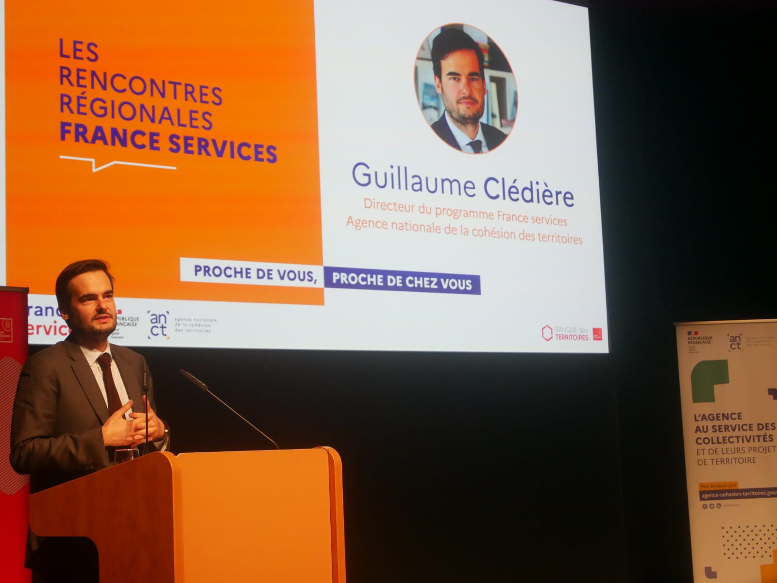 Guillaume Clédière parle à une tribune avec, derrière lui, un grand écranqui annononce l'événement  et présente sa fonction. 