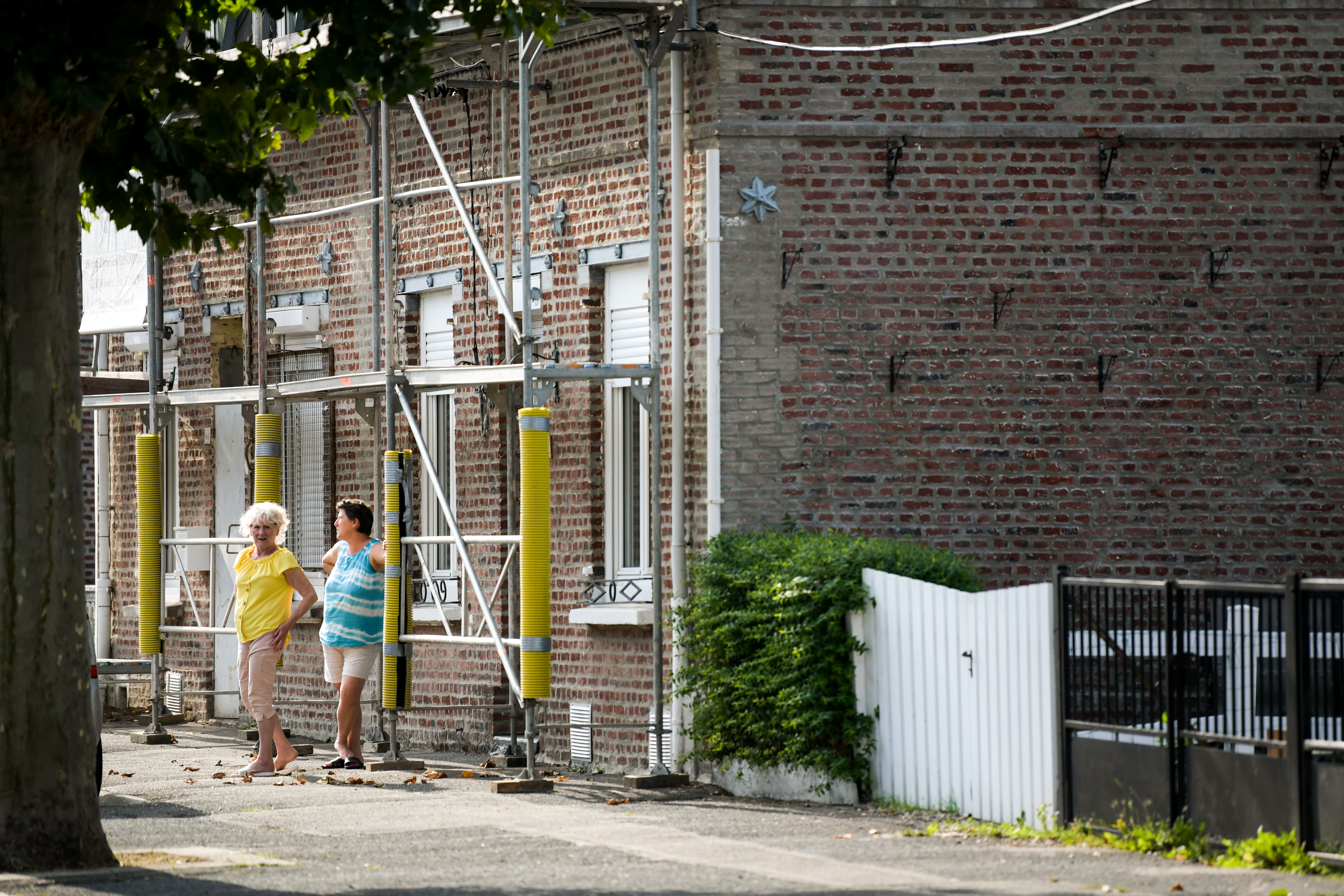 Deux femmes discutent devant une maison en brique, qui a des échaffaudages pour des travaux derénovation.