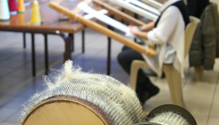 Vue sur des bobines de laine à filer, au 1er plan, et une artisan avec un métier à tisser traditionnel en bois, en arrière plan.