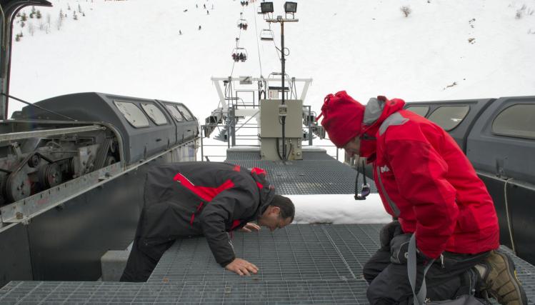 Deux techniciens en rouge vérifient un élément d'une remontée mécanique, au départ ; en arrière-plan la remontée surplombe la montagne enneigée.