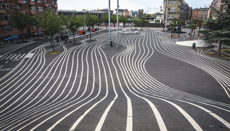 Photo d'une place d'une ville : sur le sol sont tracées de grandes lignes blanches qui évitent les obstacles du mobilier urbain et guident les déplacements des piétons.