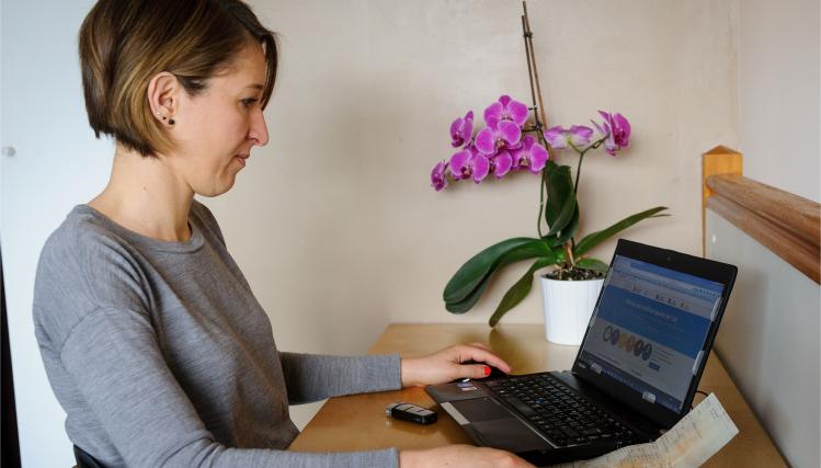 Une femme est à son bureau, chez elle, devant son ordinateur. Elle est prise de profil. Il y a une orchidée violette sur le bureau.