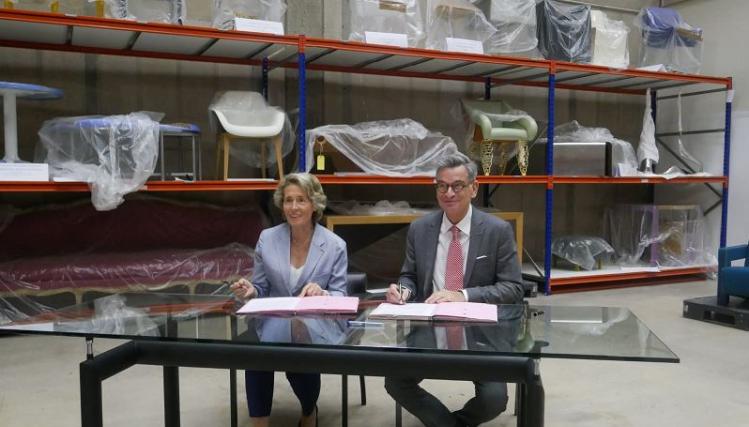 Caroline Cayeux et Hervé Lemoine signent une convenion sur une table installée au millieu d'une réserve avec du mobilier enveloppé de papier bulle, sur des étagères de stockage.