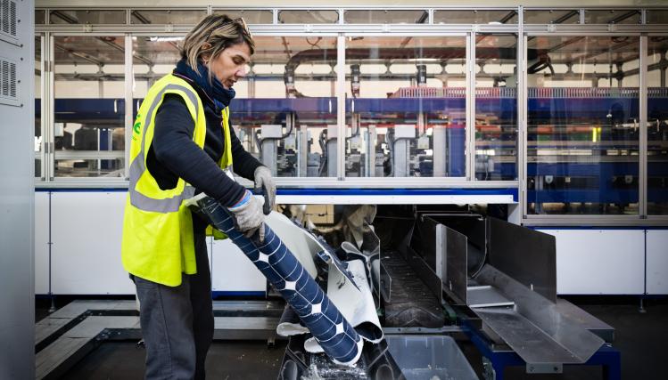 Une femme avec un gilet jaune manipule un rouleau de vinyle bleu dans une usine de recyclage.