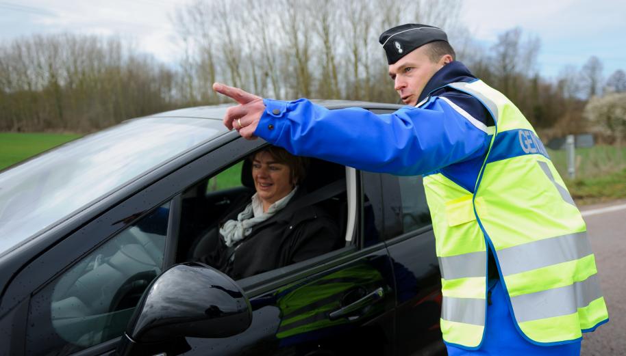 Un gendarme avec un gilet fluo renseigne une autmobiliste sur une route de campagne.