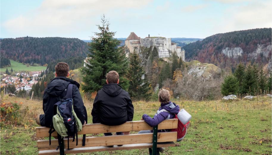 Dans un paysage de moyenne montagne, trois randonneurs assis sur un banc, et de dos, regarde un paysage avec un vieux château sur un piton rocheux, et un village en contrebas.